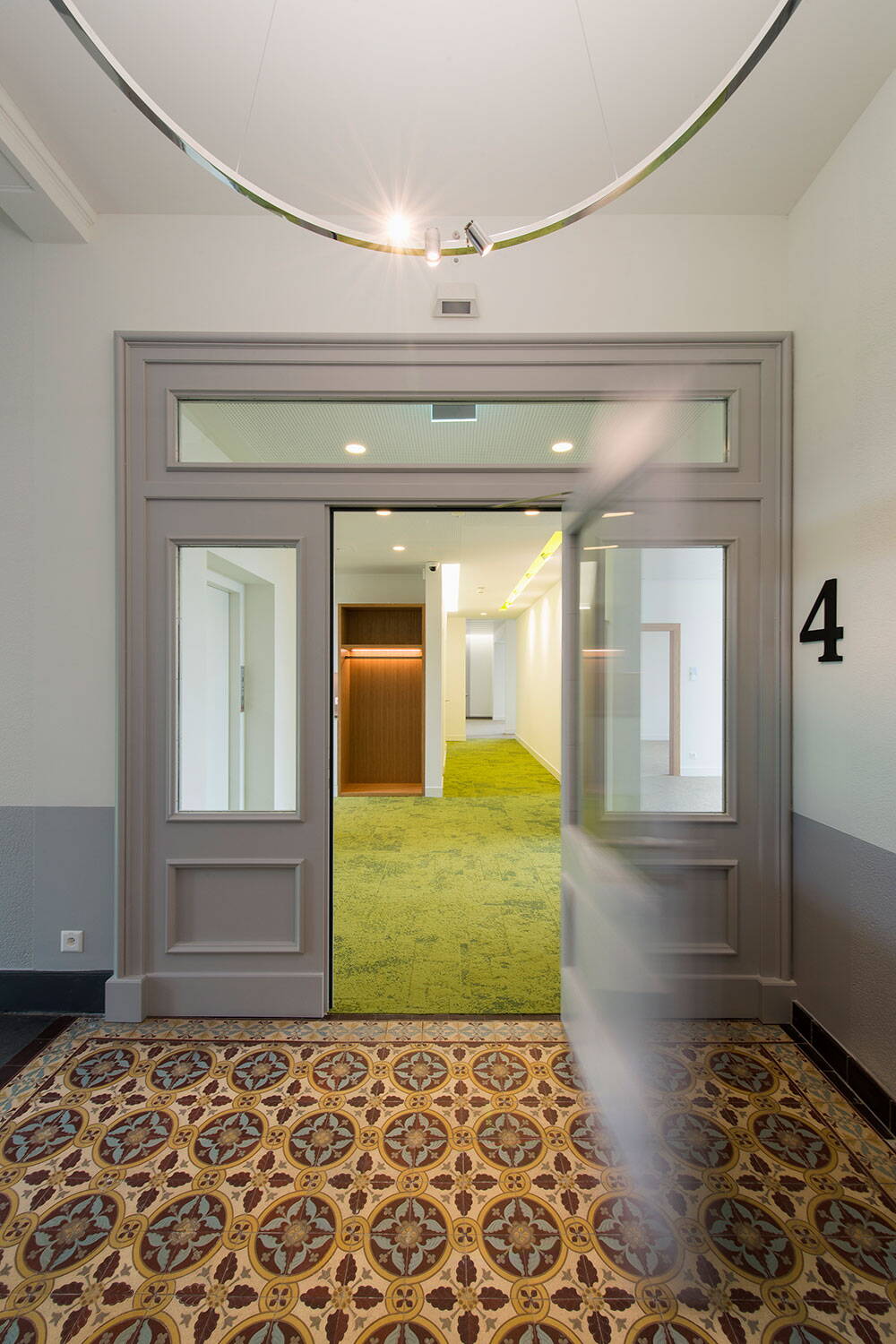 Sicht aus Treppenhaus einer renovierten Geschäftsliegenschaft durch eine offene Holztüre in einen Büroraum. Kunstvolle Bodenplatten und grüner Teppich.