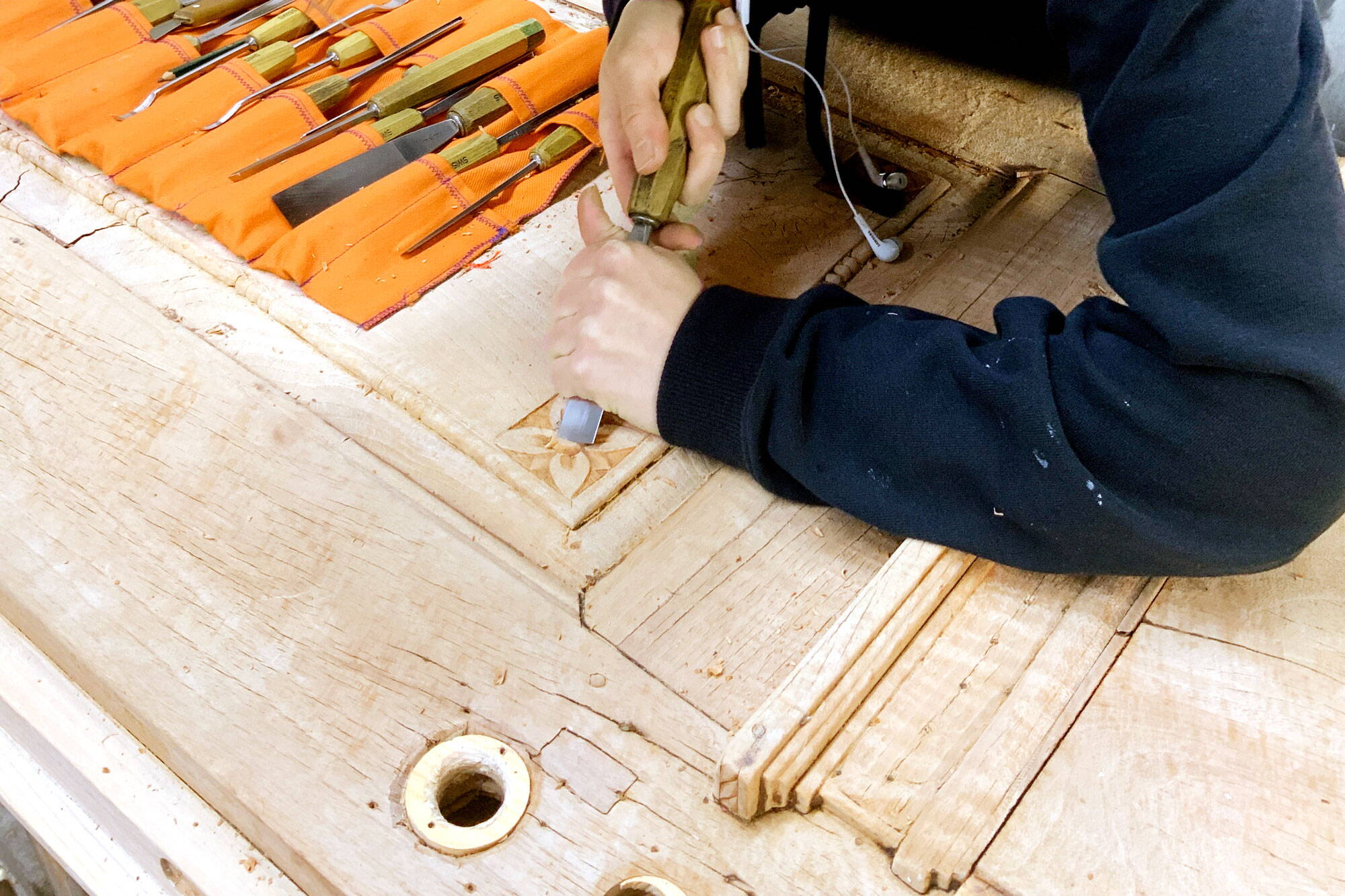 Nahaufnahme von Händen, die mit einem Stechbeutel die Verzierung einer antiken Holztüre bearbeiten. Weitere Werkzeuge im Hintergrund.