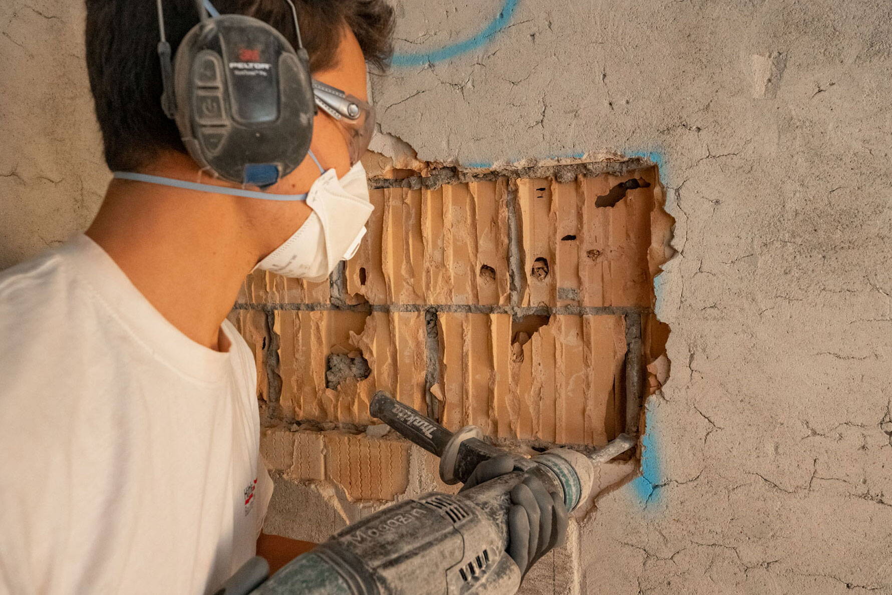 Ein Mann mit Schutzmaske und Ohrschutz bearbeitet eine Wand mit dem Schlagbohrer.