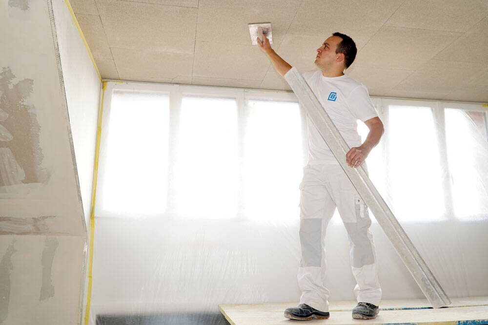 Ein Gipser in weisser Arbeitskleidung verputzt eine Decke in einem mit Folie ausgekleideten Raum. 